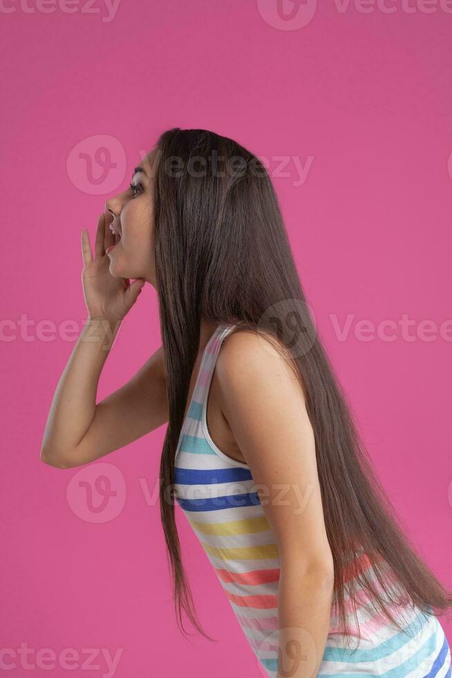 Brunette woman with long hair, dressed in colorful striped shirt, posing against pink studio background. Sincere emotions. Close-up. photo