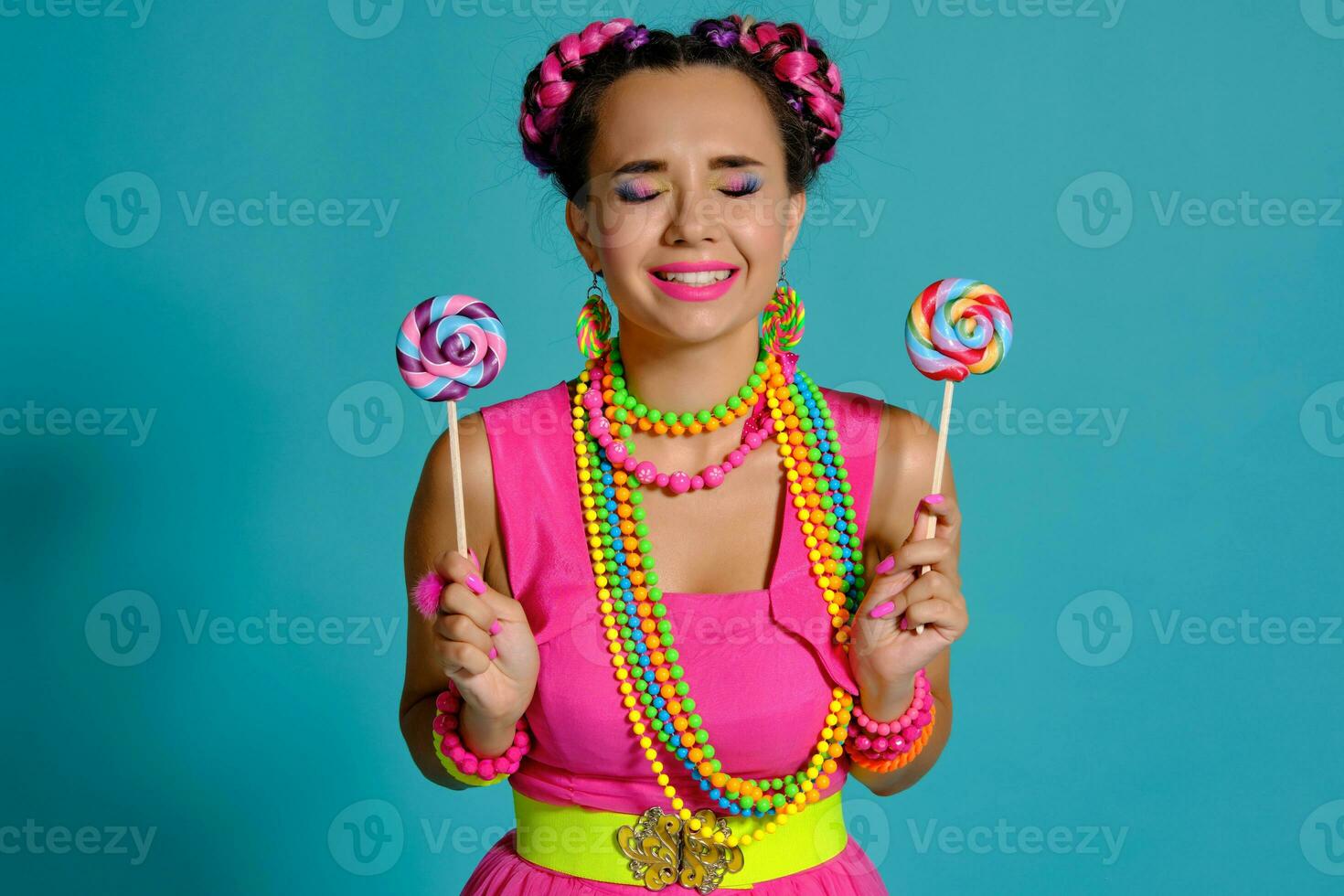 Lovely girl with a multi-colored braids hairstyle and bright make-up, posing in studio against a blue background, holding a lollipop in her hand. photo