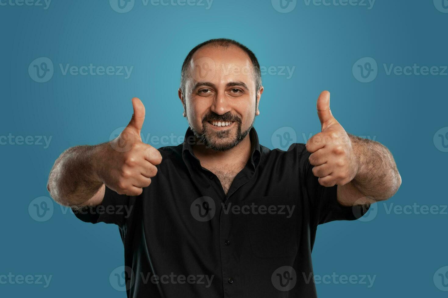 de cerca retrato de un moreno de edad mediana hombre con barba, vestido en un negro camiseta y posando en contra un azul antecedentes. foto