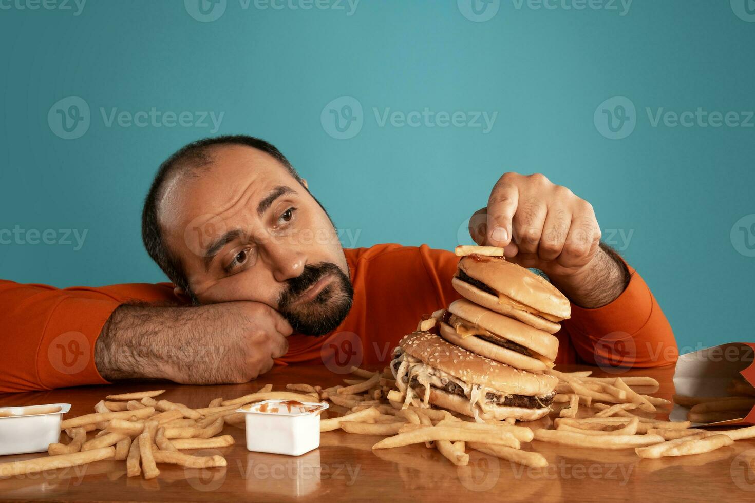 Close-up portrait of a middle-aged man with beard, dressed in a red turtleneck, posing with burgers and french fries. Blue background. Fast food. photo