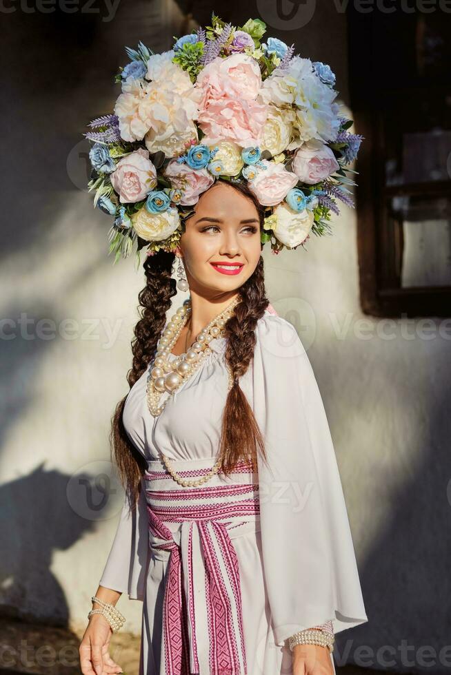 Brunette girl in a white ukrainian authentic national costume and a wreath of flowers is posing against a white hut. Close-up. photo