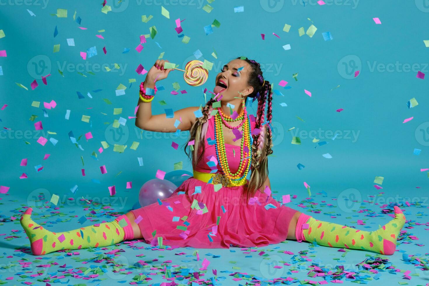 Lovely girl with a multi-colored braids hairstyle and bright make-up, posing in studio with lollipop, air balloons and confetti against a blue background. photo