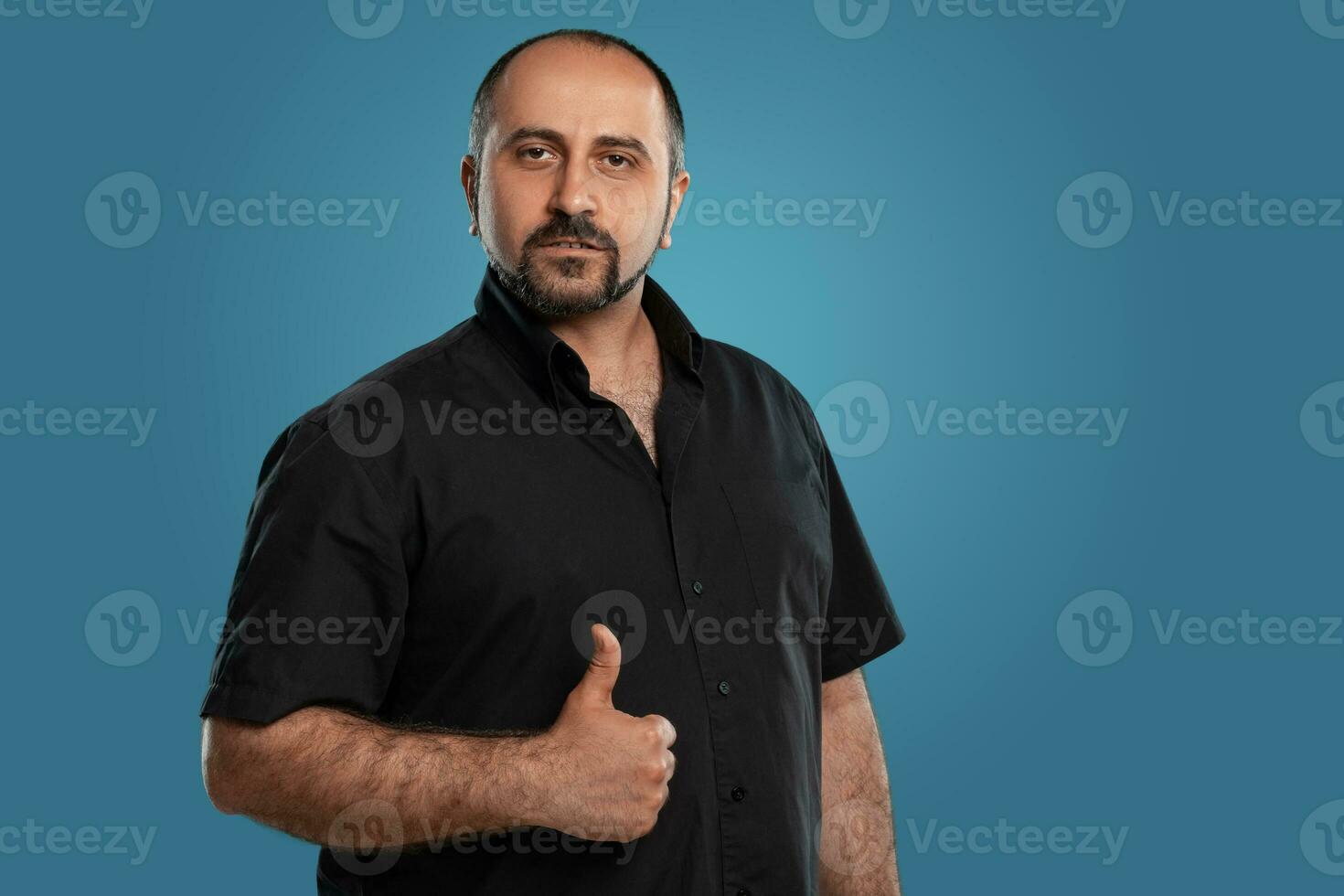 Close-up portrait of a brunet middle-aged man with beard, dressed in a black t-shirt and posing against a blue background. photo