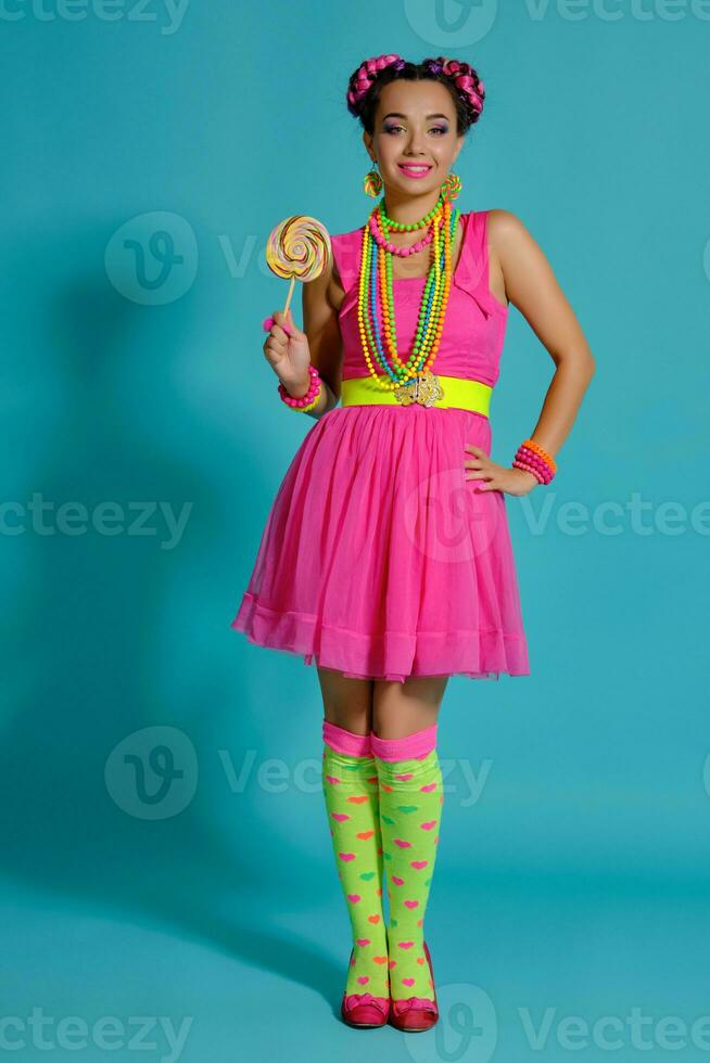 Lovely girl with a multi-colored braids hairstyle and bright make-up, posing in studio against a blue background, holding a lollipop in her hand. photo