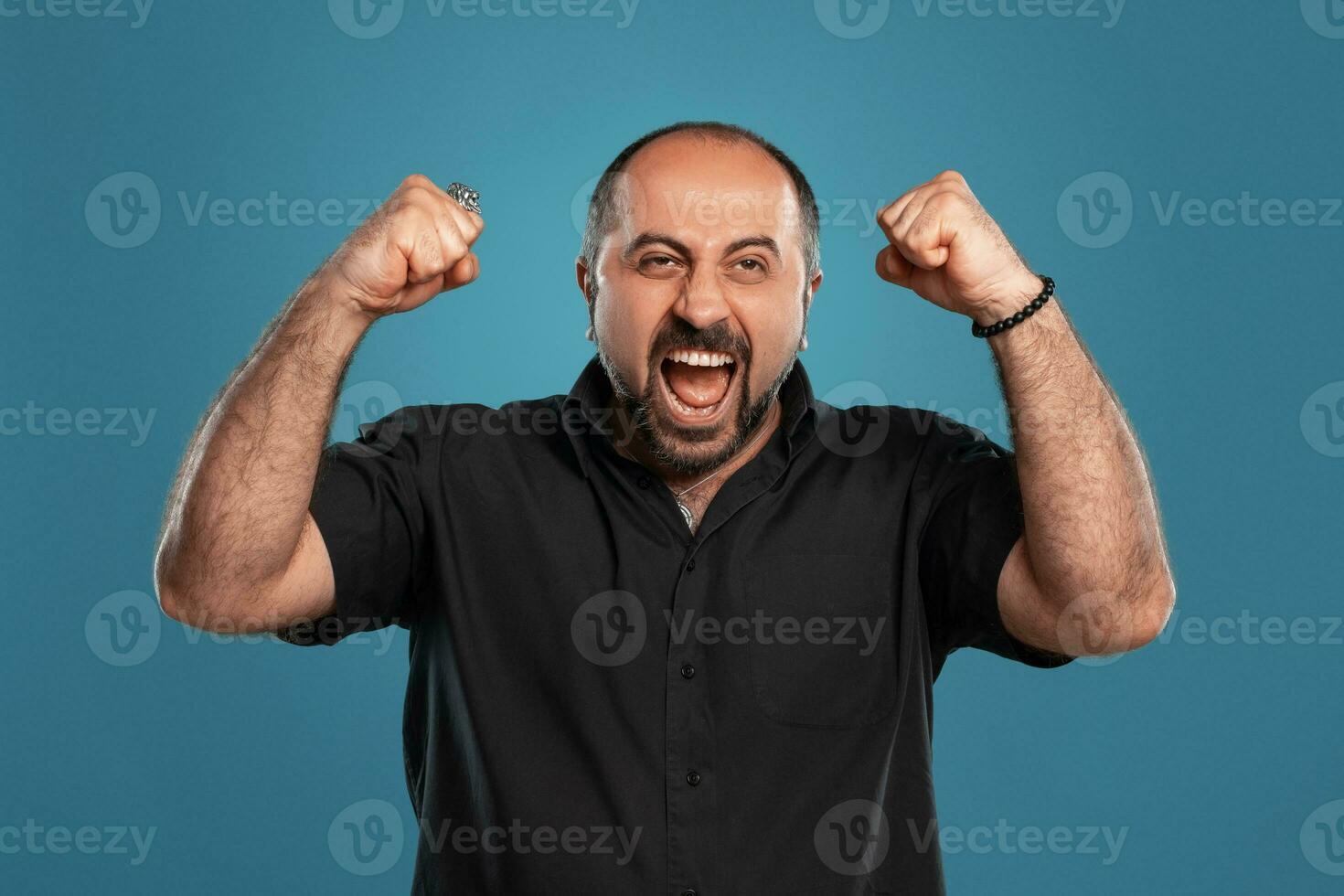 Close-up portrait of a brunet middle-aged man with beard, dressed in a black t-shirt and posing against a blue background. photo