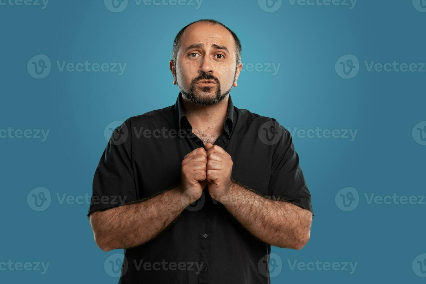 de cerca retrato de un moreno de edad mediana hombre con barba, vestido en un negro camiseta y posando en contra un azul antecedentes. foto