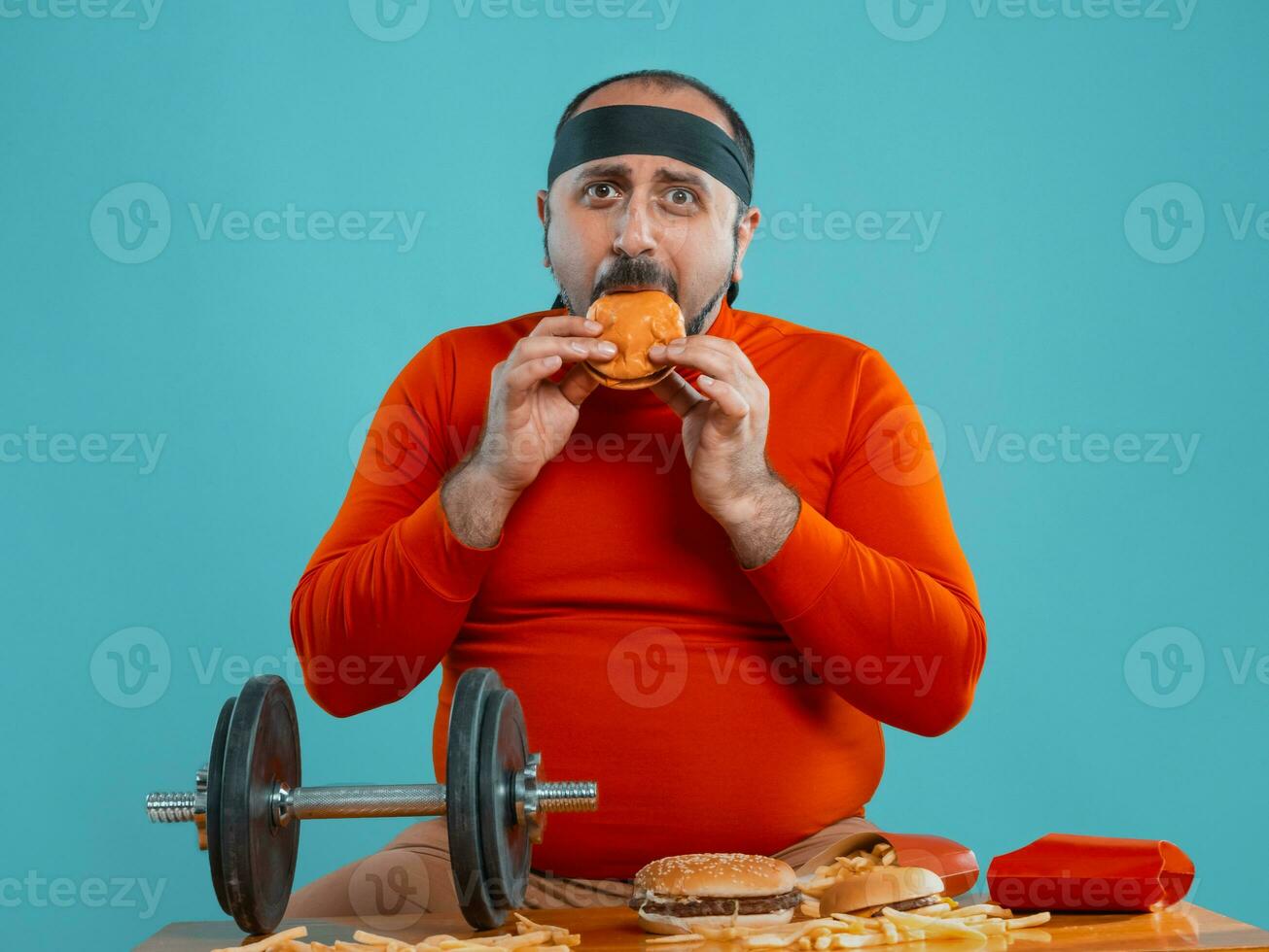 Middle-aged man with beard, dressed in a red turtleneck, headband, posing with burgers and french fries. Blue background. Close-up. Fast food. photo