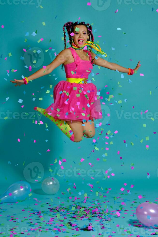 encantador niña con un multicolor trenzas peinado y brillante constituir, posando en estudio con aire globos y papel picado en contra un azul antecedentes. foto