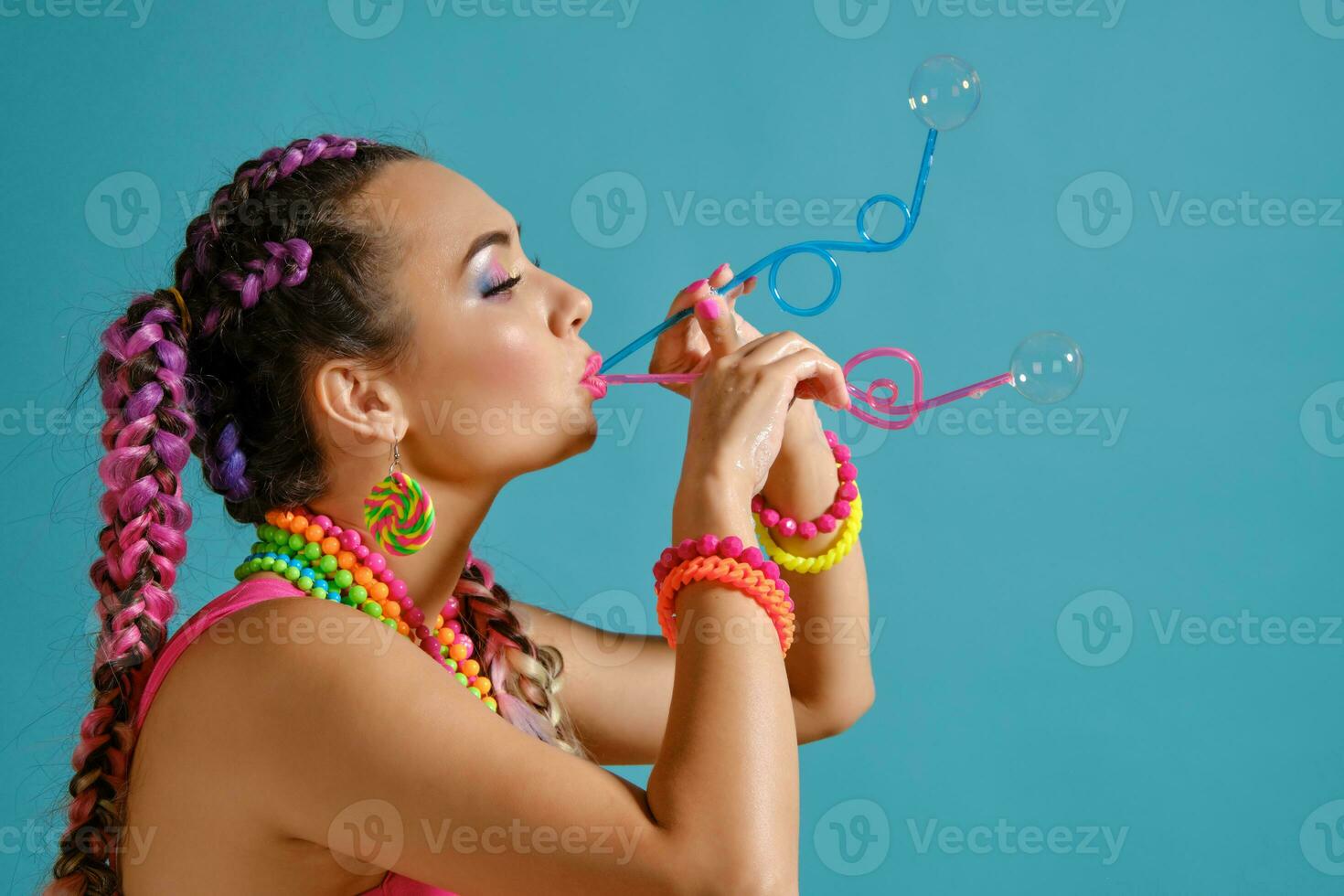 Lovely girl with a multi-colored braids hairstyle and bright make-up, is blowing bubbles using tubules, posing in studio against a blue background. photo