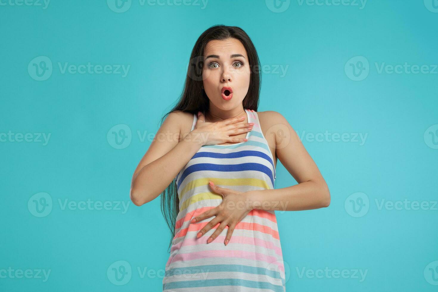 Brunette woman with long hair, dressed in colorful striped shirt, posing against blue studio background. Sincere emotions. Close-up. photo