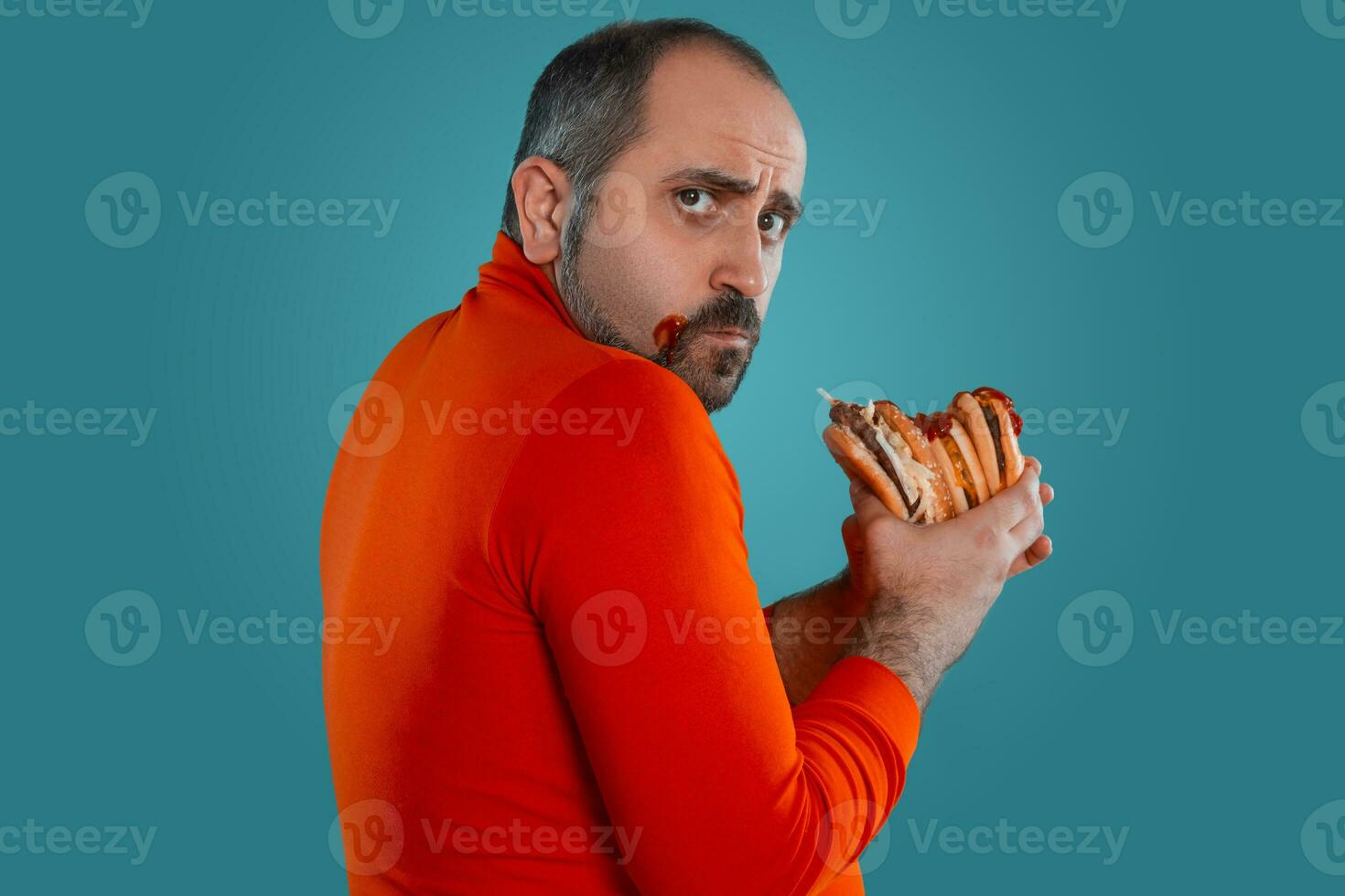 de cerca retrato de un de edad mediana hombre con barba, vestido en un rojo suéter tipo con cuello de tortuga, posando con hamburguesas en contra un azul antecedentes. rápido alimento. foto