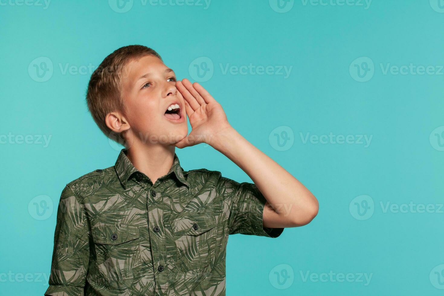 Close-up portrait of a blonde teenage boy in a green shirt with palm print posing against a blue studio background. Concept of sincere emotions. photo