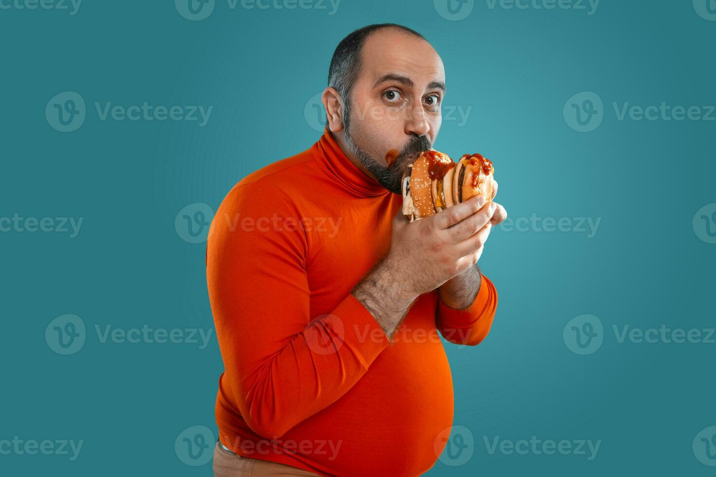 Close-up portrait of a middle-aged man with beard, dressed in a red turtleneck, posing with burgers against a blue background. Fast food. photo