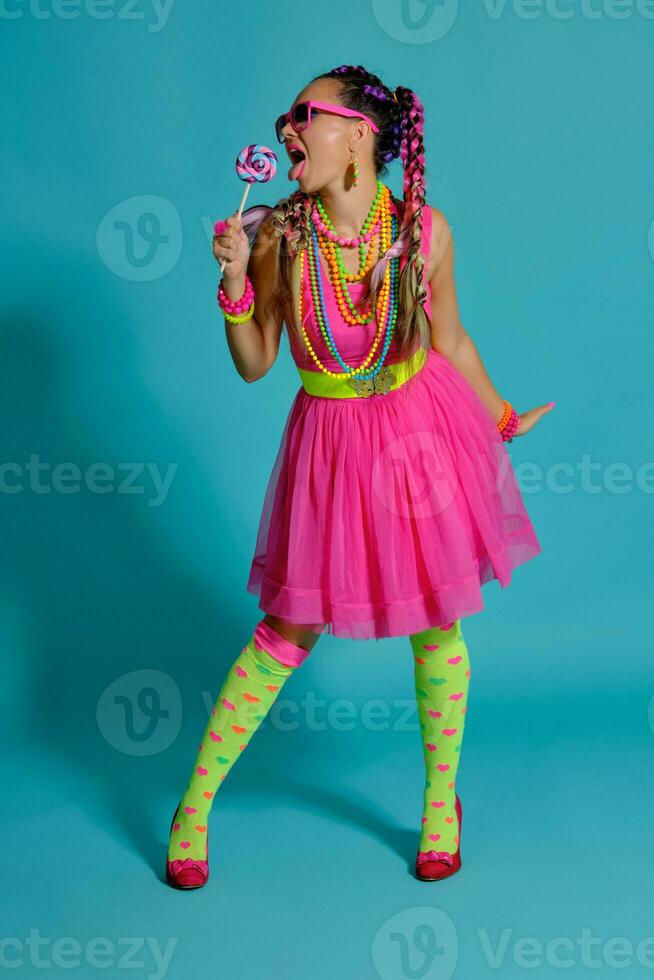 Lovely girl with a multi-colored braids hairstyle and bright make-up, posing in studio against a blue background, holding a lollipop in her hand. photo