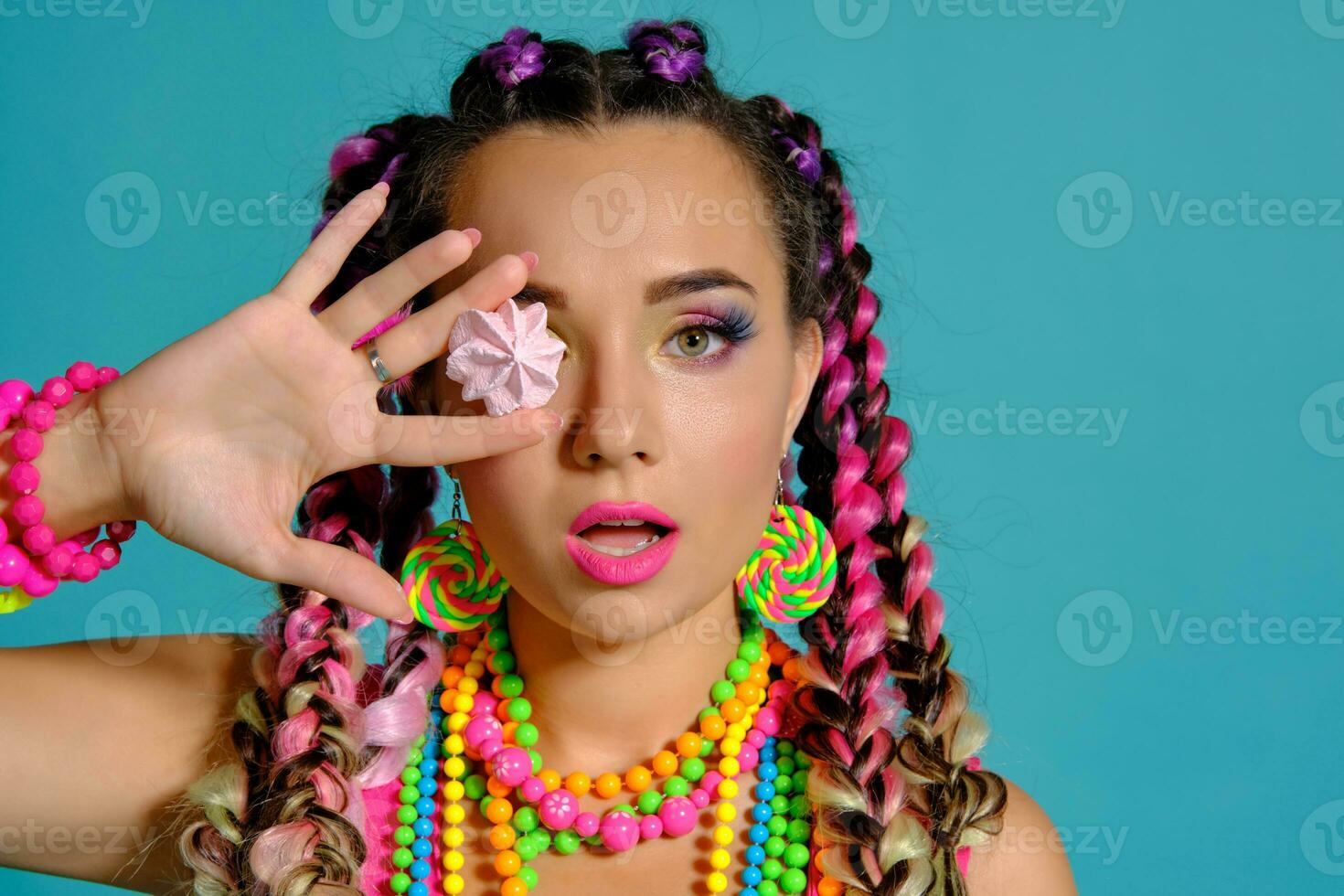 Lovely girl with a multi-colored braids hairstyle and bright make-up, posing in studio against a blue background, holding marshmallow in her hands. photo