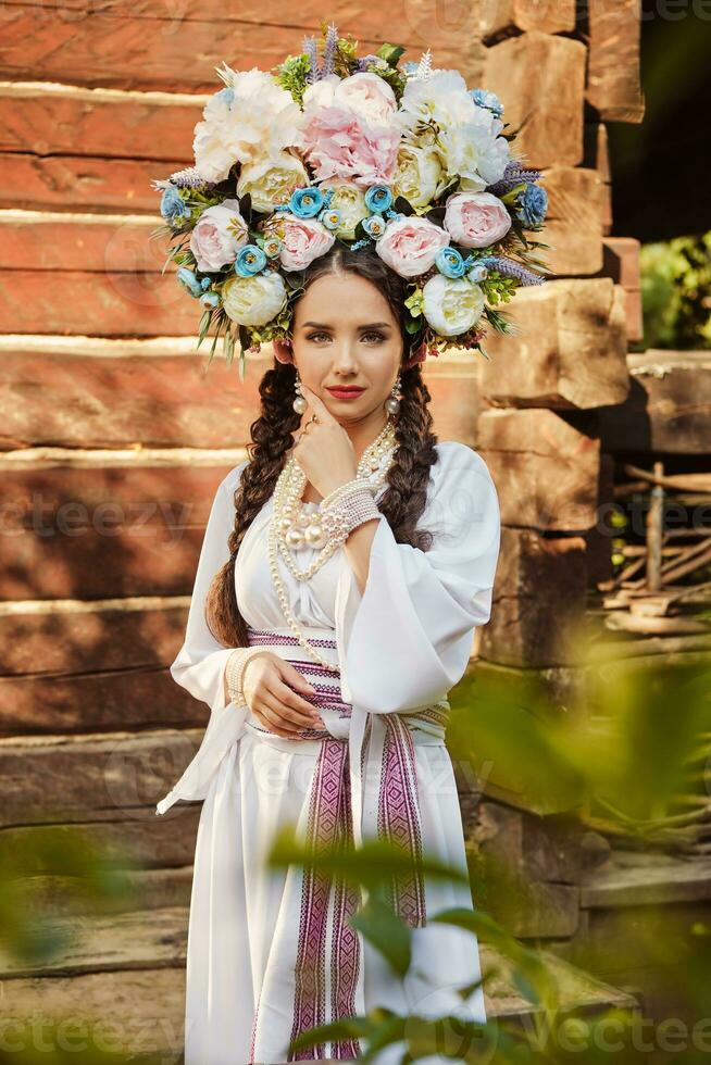 morena niña en un blanco ucranio auténtico nacional disfraz y un guirnalda de flores es posando en contra un de madera casa. de cerca. foto