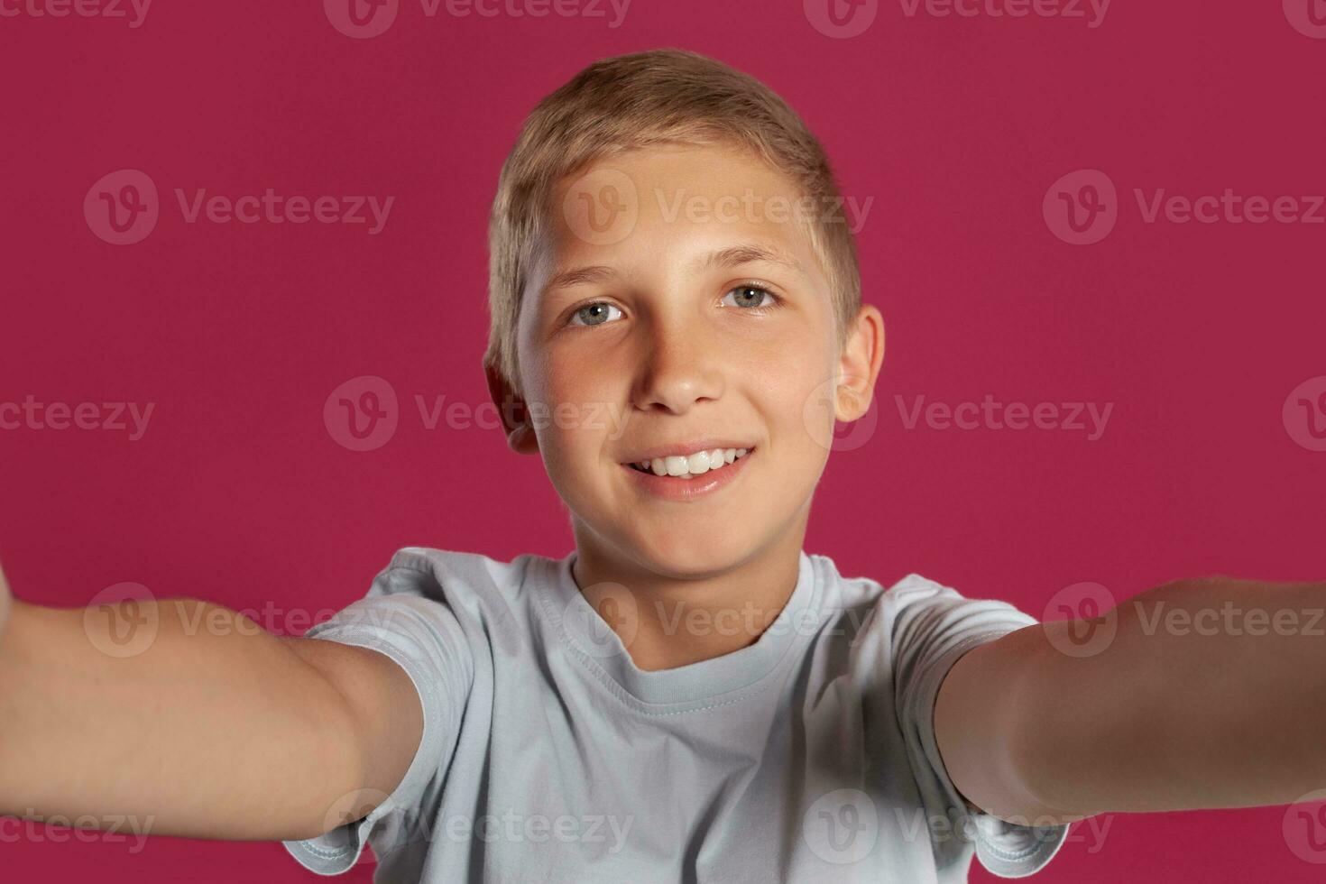 Close-up portrait of a blonde teenage boy in a white t-shirt posing against a pink studio background. Concept of sincere emotions. photo