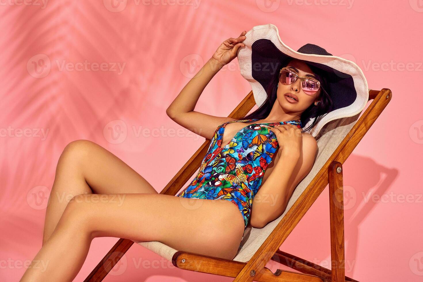 Summer lifestyle fashion portrait of young stunning woman on a lounge chair, pink background, studio shot. Wearing stylish sunglasses, bikini. photo