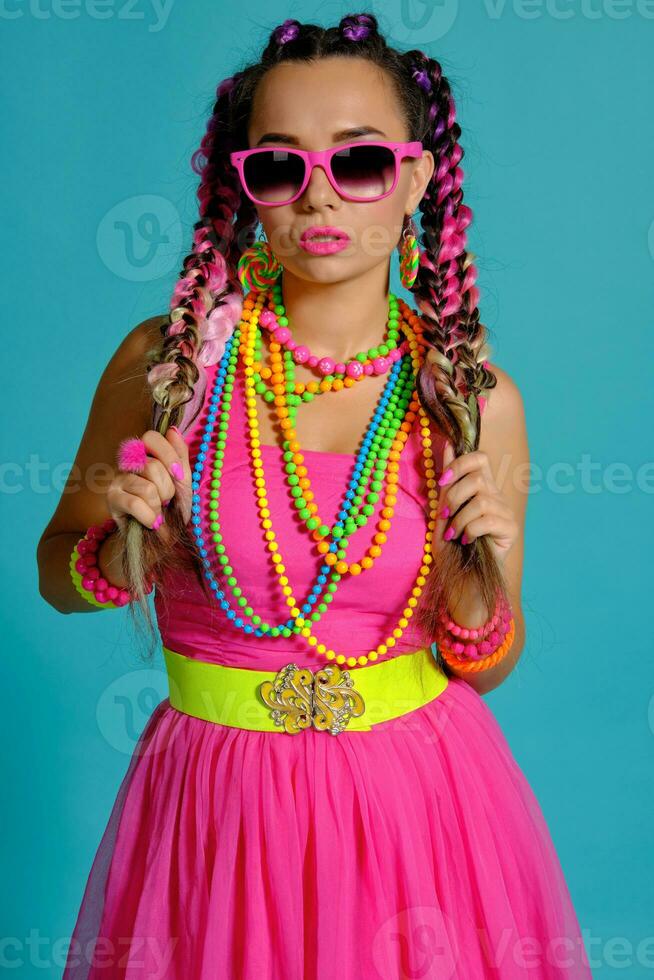 encantador niña con un multicolor trenzas peinado y brillante constituir, posando en estudio en contra un azul fondo, participación un pirulí en su mano. foto