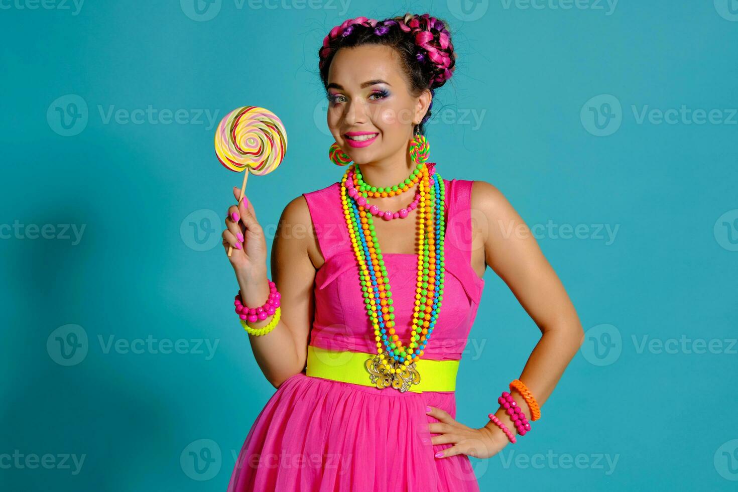 encantador niña con un multicolor trenzas peinado y brillante constituir, posando en estudio en contra un azul fondo, participación un pirulí en su mano. foto