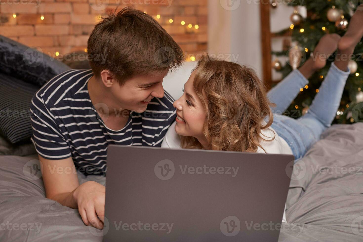 Smiling young couple man and woman lying in bed with laptop and looking at each other photo