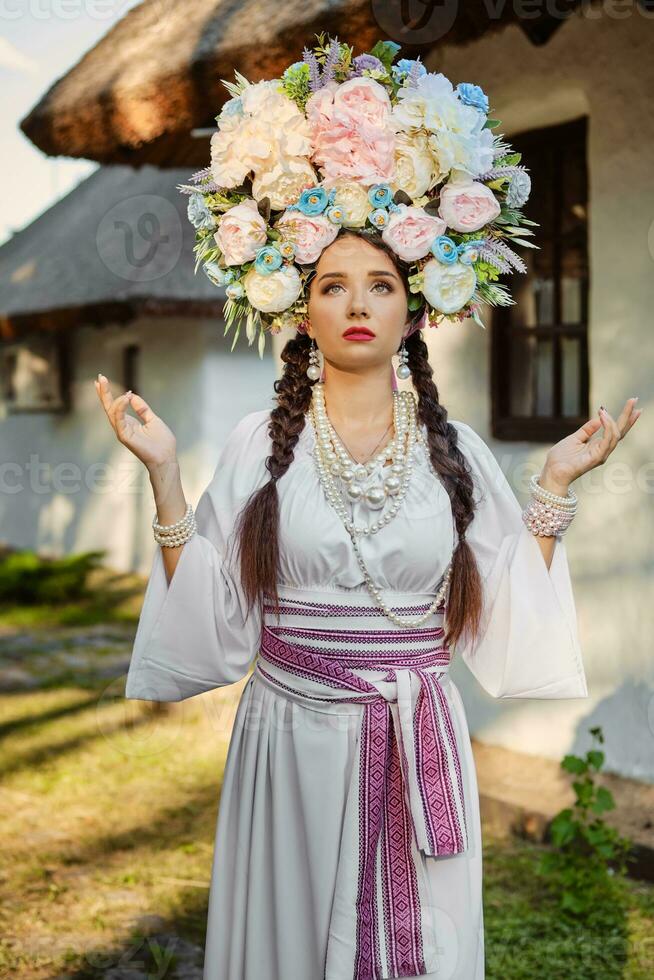 Brunette girl in a white ukrainian authentic national costume and a wreath of flowers is posing against a white hut. Close-up. photo