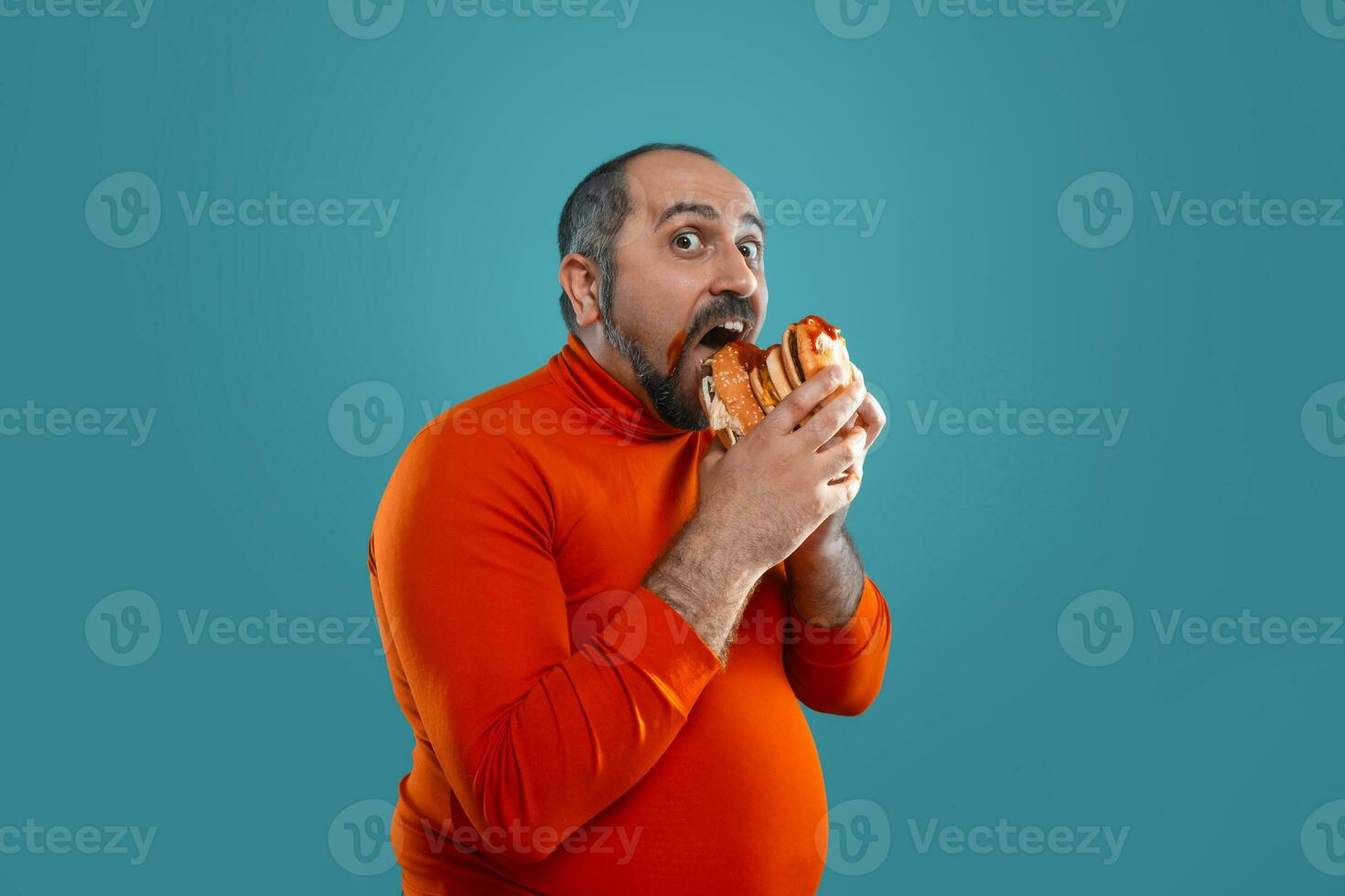 Close-up portrait of a middle-aged man with beard, dressed in a red turtleneck, posing with burgers against a blue background. Fast food. photo