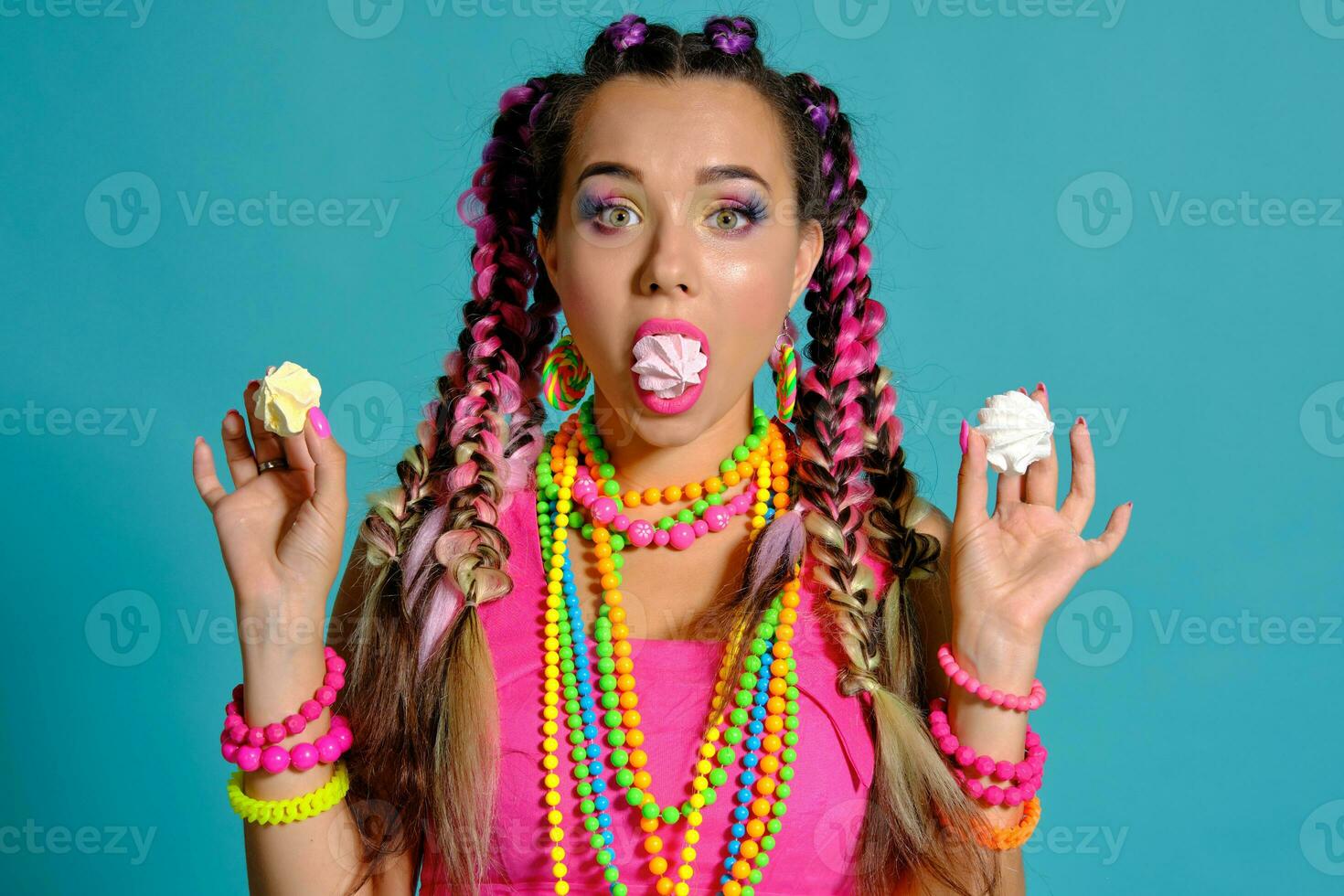 Lovely girl with a multi-colored braids hairstyle and bright make-up, posing in studio against a blue background, holding marshmallow in her hands. photo