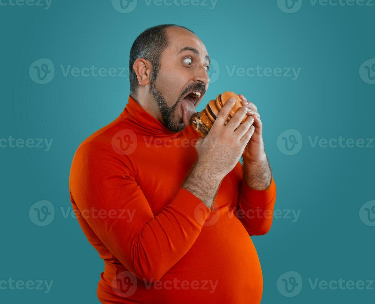 Close-up portrait of a middle-aged man with beard, dressed in a red turtleneck, posing with burgers against a blue background. Fast food. photo