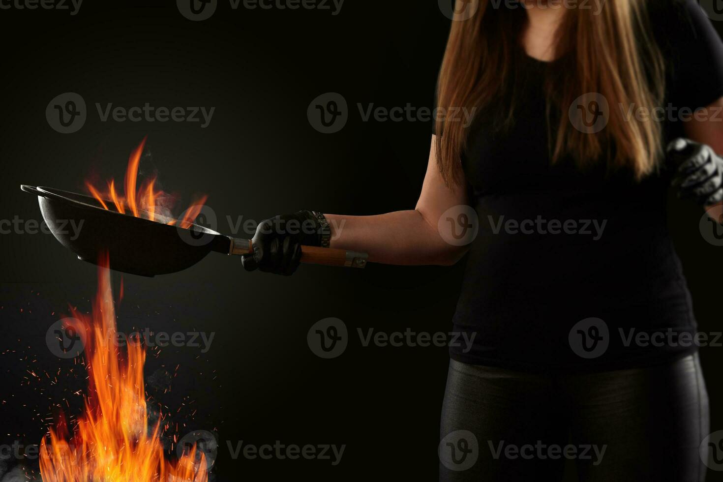 Brunette lady with tattooed hands, dressed in leggings and t-shirt. Holding a wok pan above fire against black background. Cooking concept. Side view photo