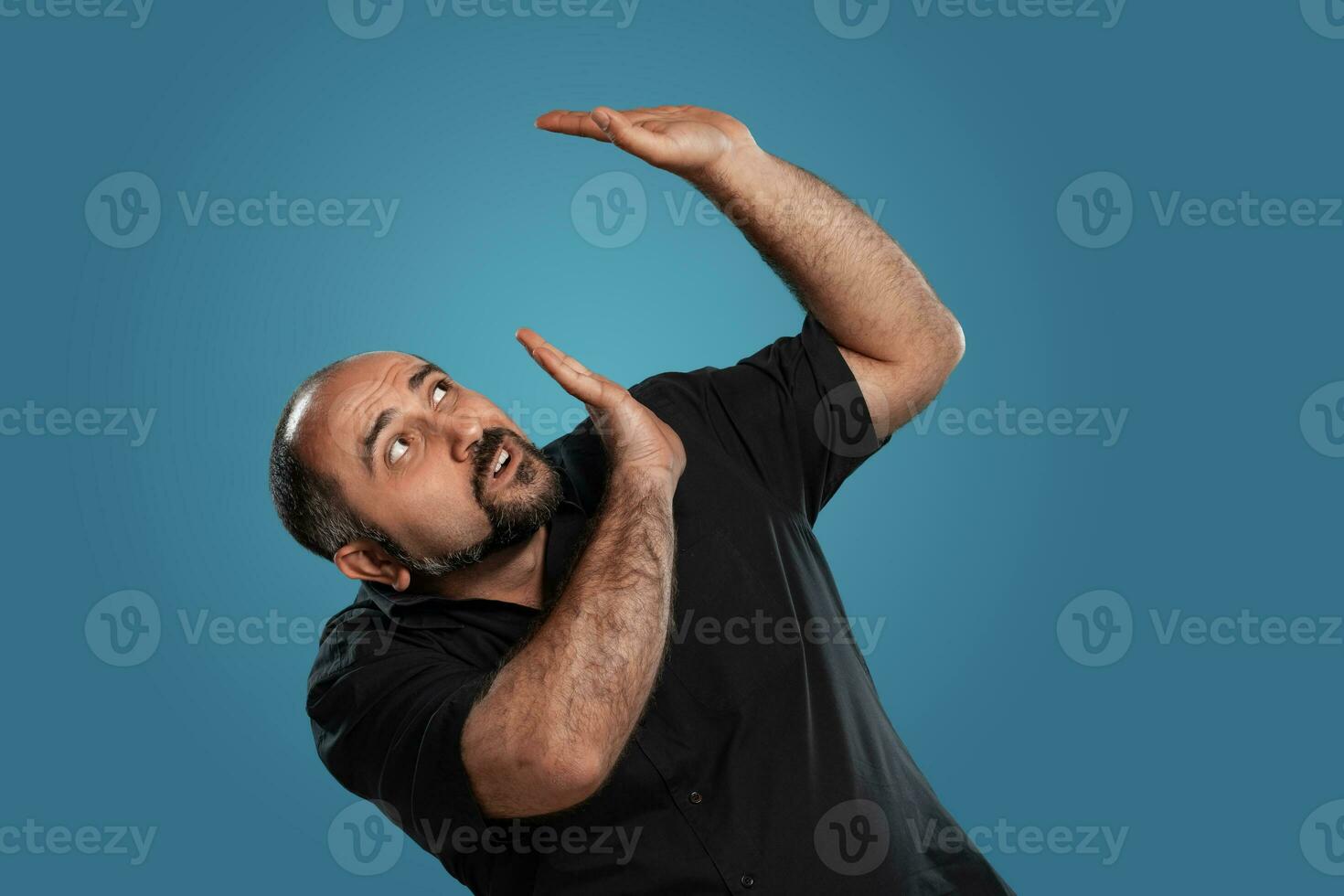 Close-up portrait of a brunet middle-aged man with beard, dressed in a black t-shirt and posing against a blue background. photo