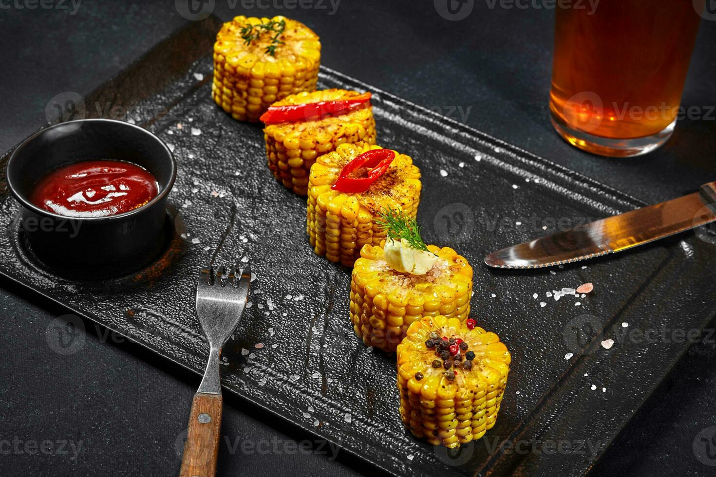 Sliced boiled corn ear with salt, chili peppers, butter and tomato sauce on slate board photo