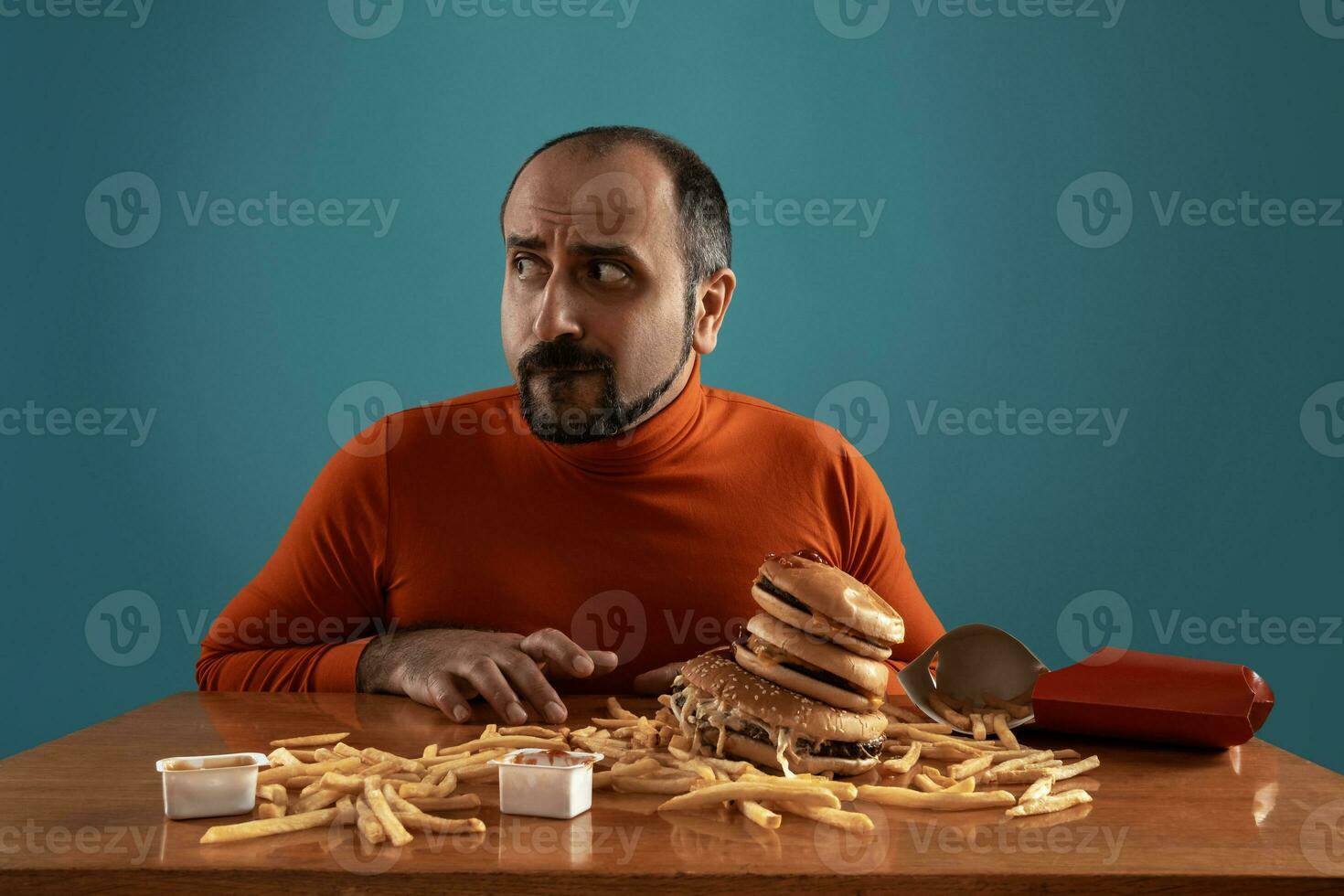 Close-up portrait of a middle-aged man with beard, dressed in a red turtleneck, posing with burgers and french fries. Blue background. Fast food. photo