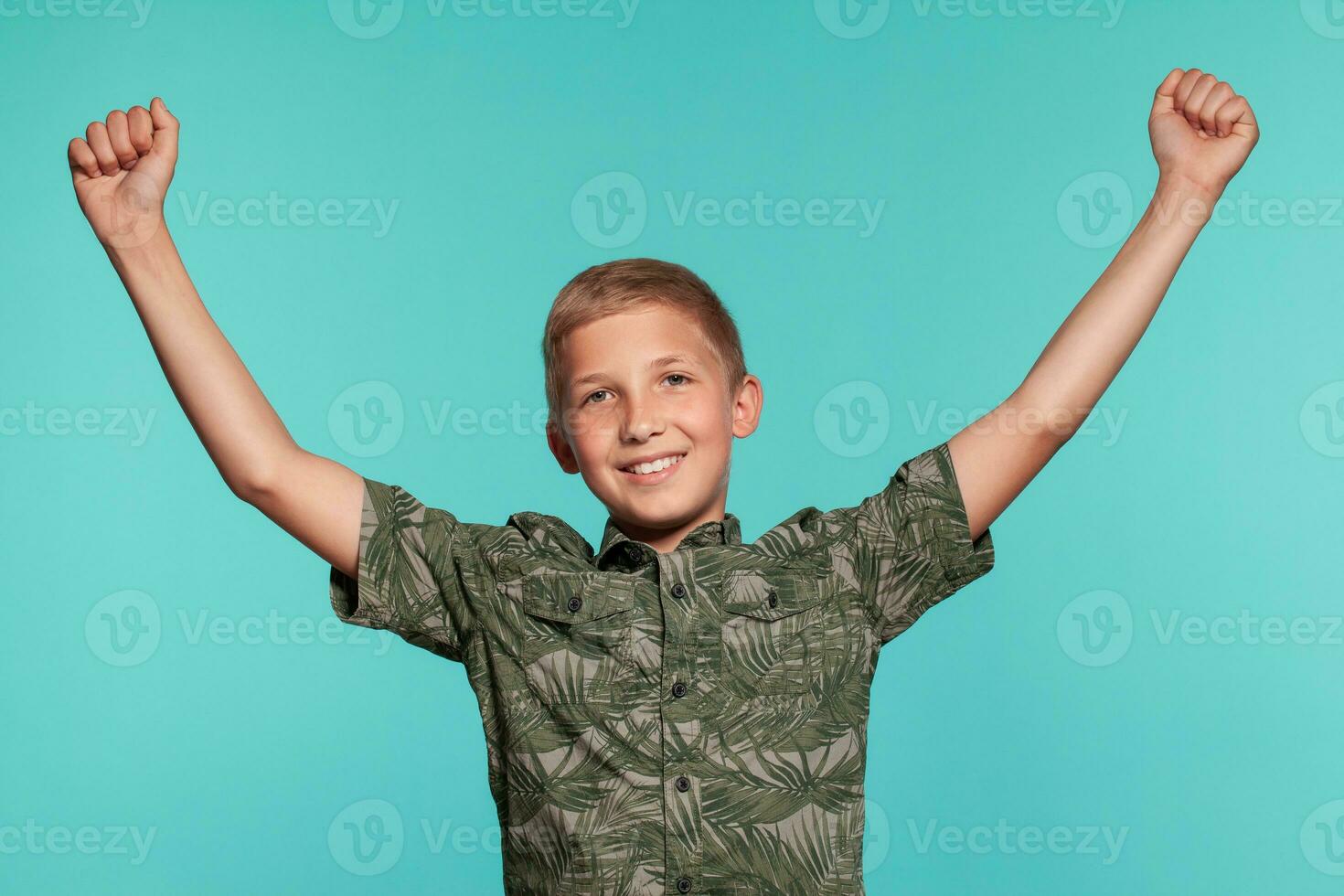 Close-up portrait of a blonde teenage boy in a green shirt with palm print posing against a blue studio background. Concept of sincere emotions. photo