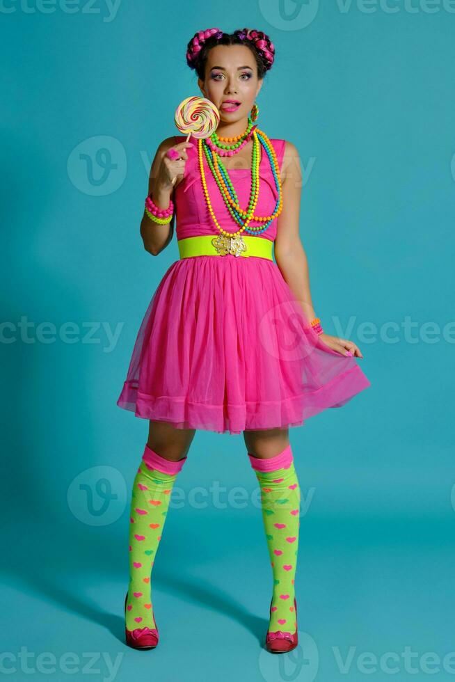 Lovely girl with a multi-colored braids hairstyle and bright make-up, posing in studio against a blue background, holding a lollipop in her hand. photo