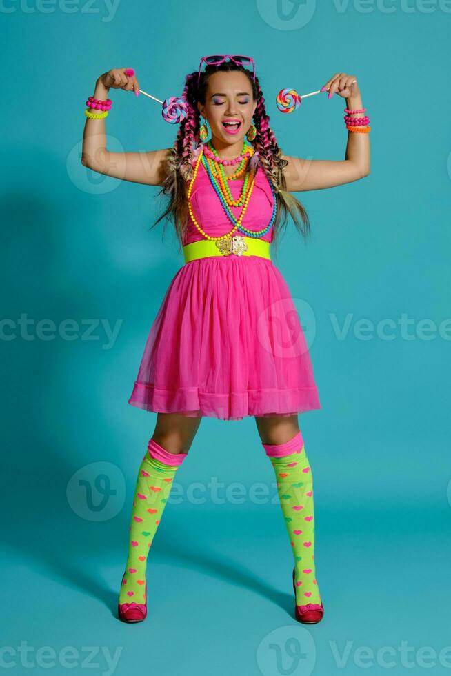 Lovely girl with a multi-colored braids hairstyle and bright make-up, posing in studio against a blue background, holding a lollipop in her hand. photo