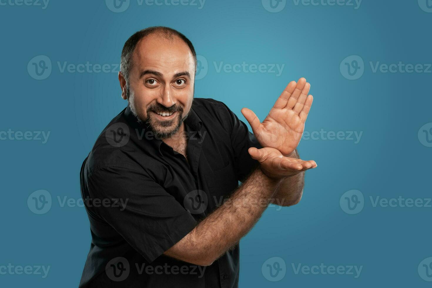 Close-up portrait of a brunet middle-aged man with beard, dressed in a black t-shirt and posing against a blue background. photo