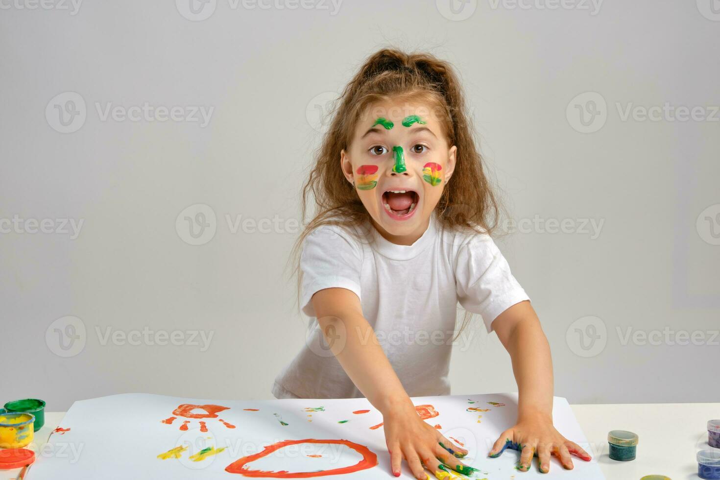 Little girl in white t-shirt sitting at table with whatman and paints on it, posing with painted face and hands. Isolated on white. Medium close-up. photo