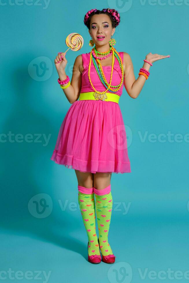 Lovely girl with a multi-colored braids hairstyle and bright make-up, posing in studio against a blue background, holding a lollipop in her hand. photo