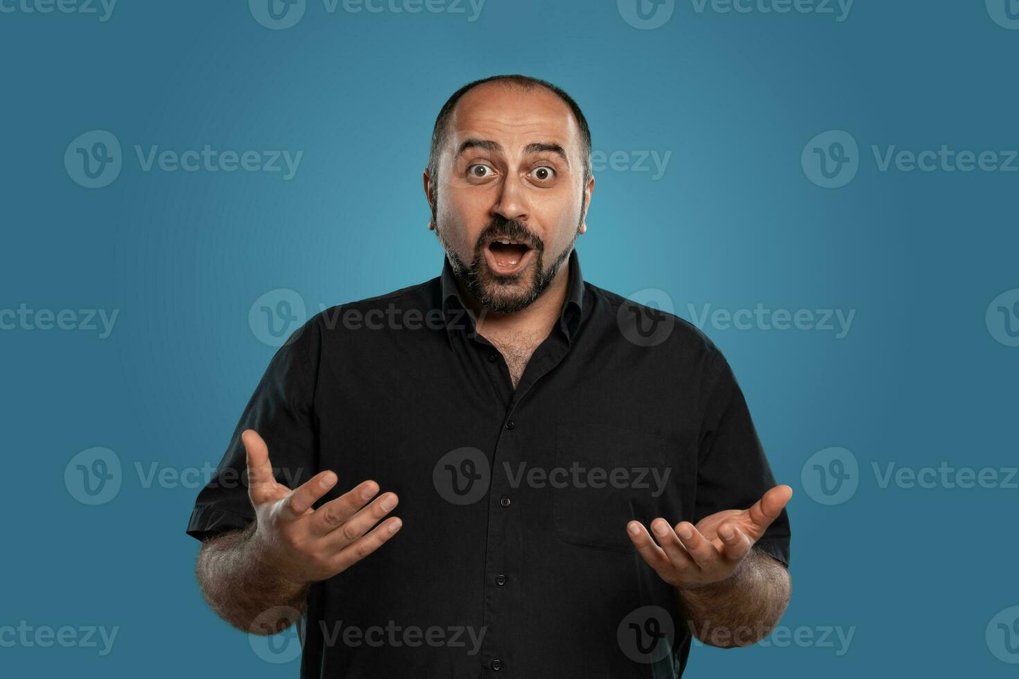 Close-up portrait of a brunet middle-aged man with beard, dressed in a black t-shirt and posing against a blue background. photo