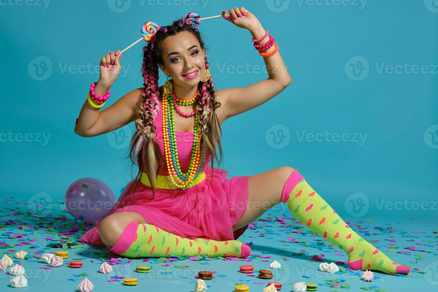 Lovely girl with a multi-colored braids hairstyle and bright make-up, posing in studio with lollipop, air balloons and confetti against a blue background. photo