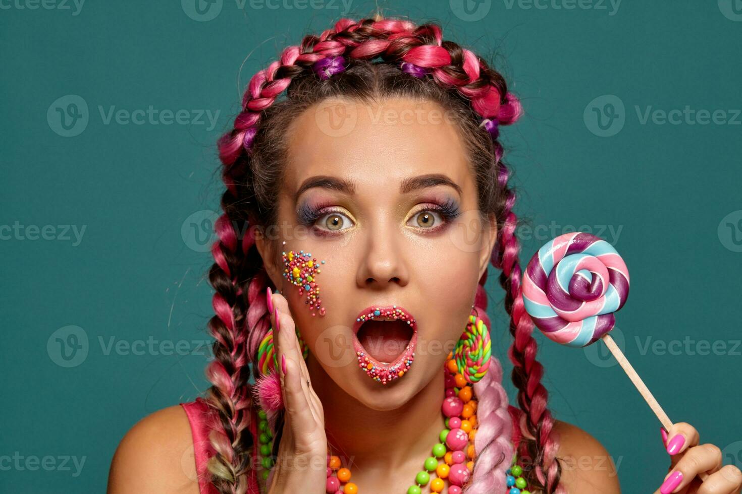 Lovely girl with a multi-colored braids hairstyle and bright make-up, posing in studio against a blue background, holding a lollipop in her hand. photo