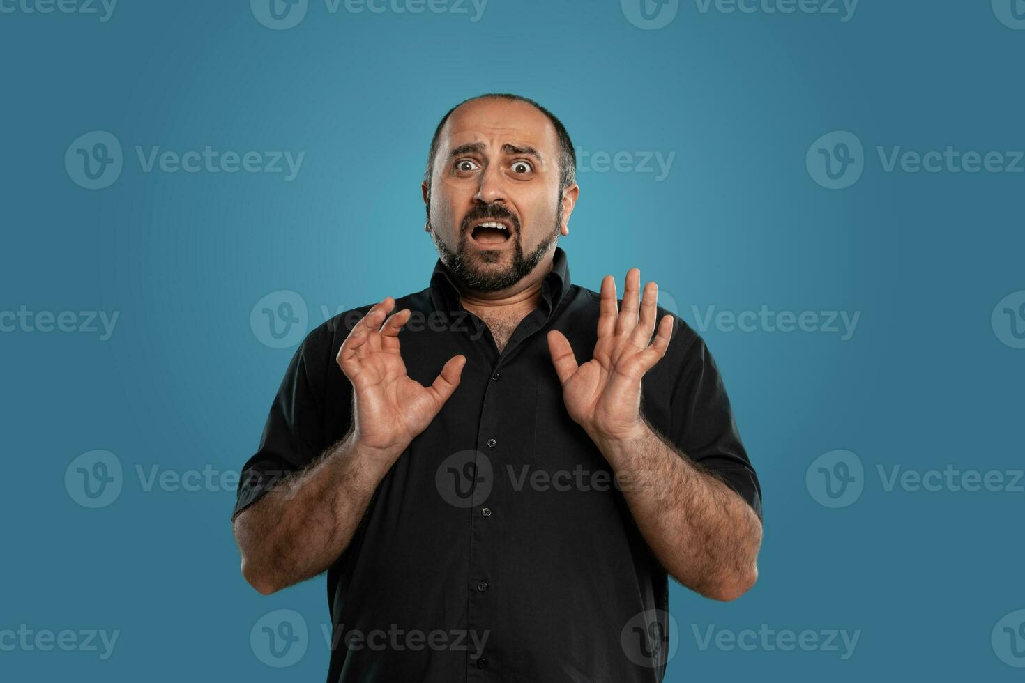 Close-up portrait of a brunet middle-aged man with beard, dressed in a black t-shirt and posing against a blue background. photo
