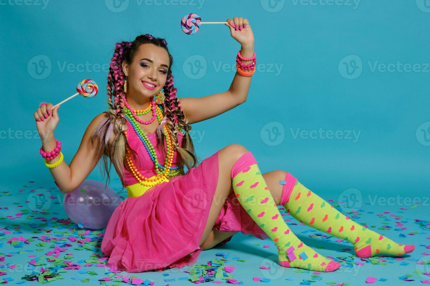 encantador niña con un multicolor trenzas peinado y brillante constituir, posando en estudio con chupete, aire globos y papel picado en contra un azul antecedentes. foto