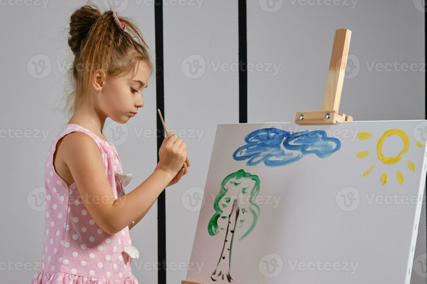 Little girl artist in a pink dress is standing behind easel and painting with brush on canvas at art studio with white walls. Medium close-up shot. photo