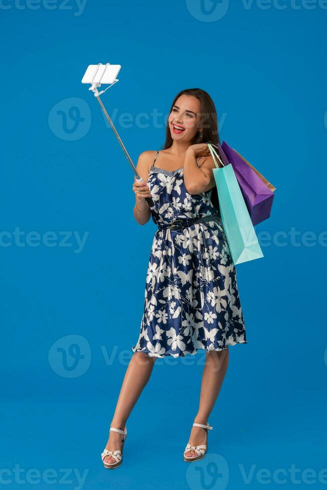 Young woman in casual clothes with shopping bags using selfie stick to take a self portrait on blue studio background with copy space photo