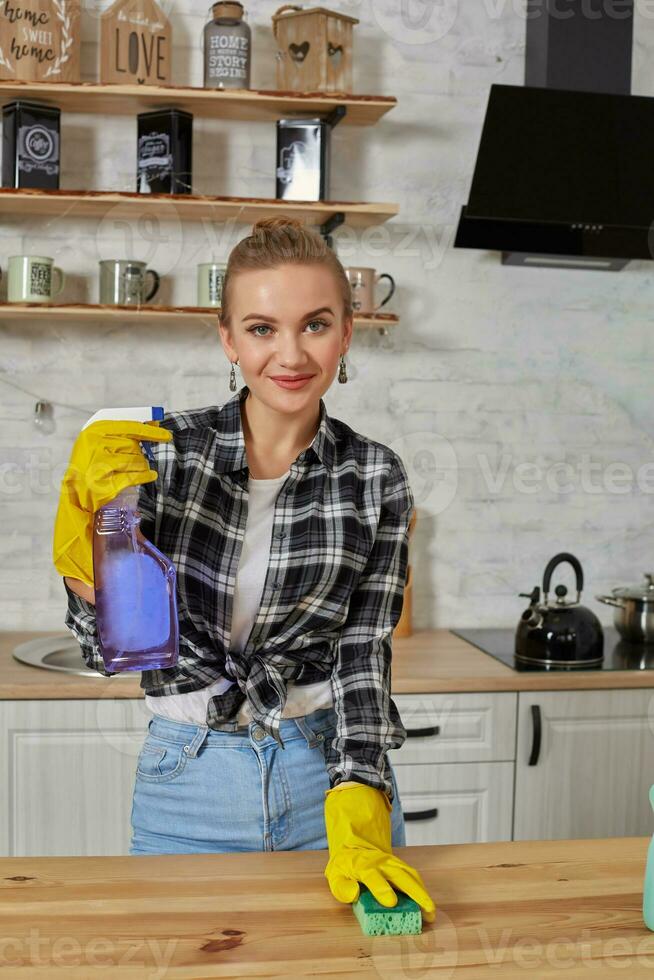 Beautiful woman in protective gloves cleaning kitchen table with rag and spray photo