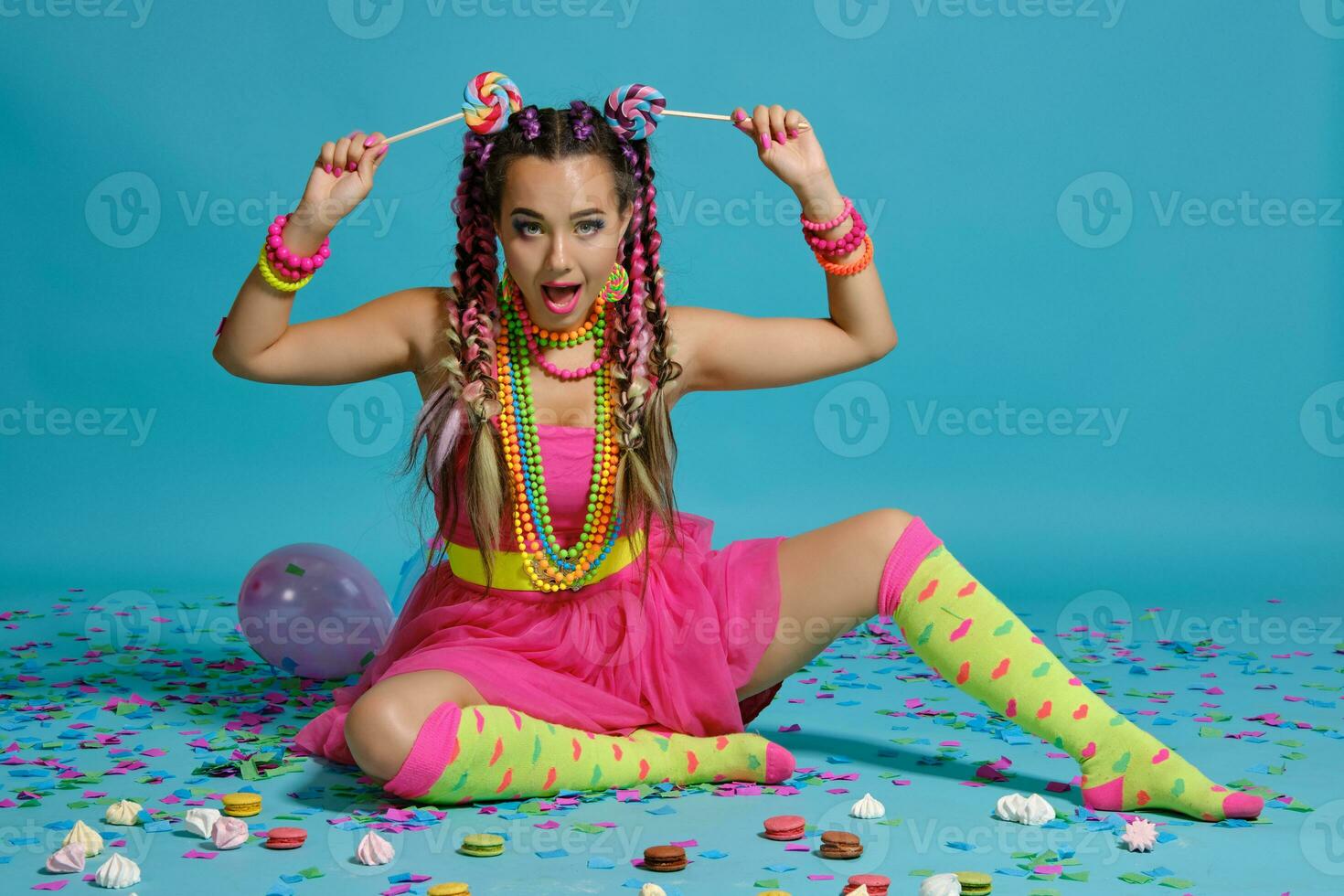 encantador niña con un multicolor trenzas peinado y brillante constituir, posando en estudio con chupete, aire globos y papel picado en contra un azul antecedentes. foto