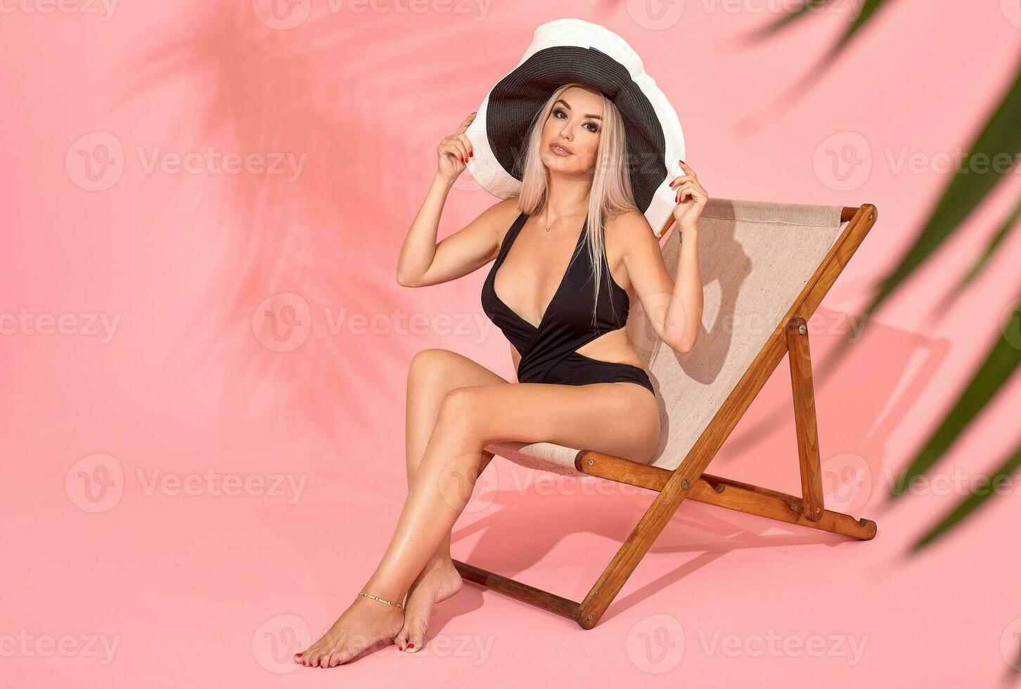 Young woman in black swimsuit relaxing on chaise longue, studio shot. Blurred palm tree leaves in foreground. photo