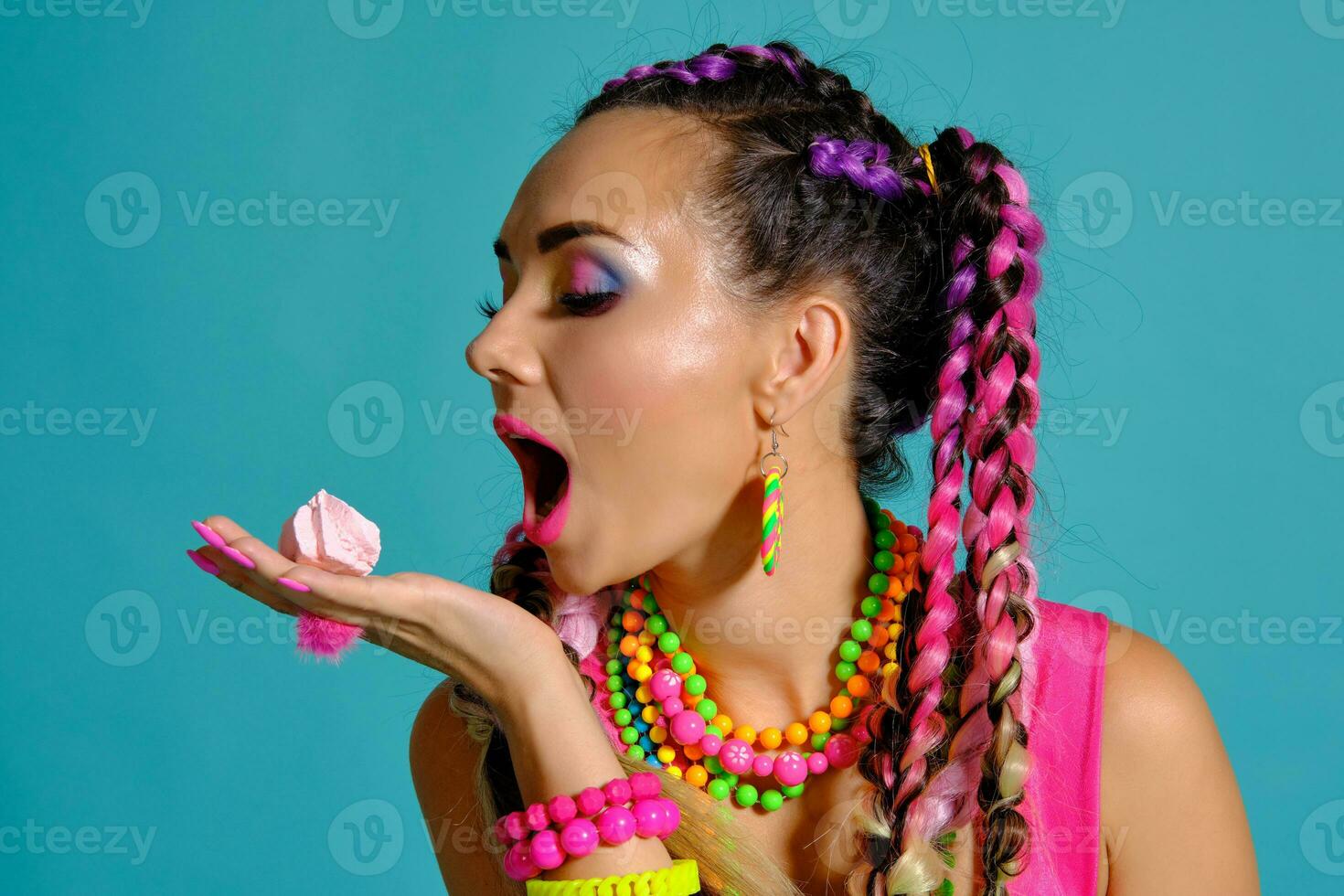 Lovely girl with a multi-colored braids hairstyle and bright make-up, posing in studio against a blue background, holding marshmallow in her hands. photo